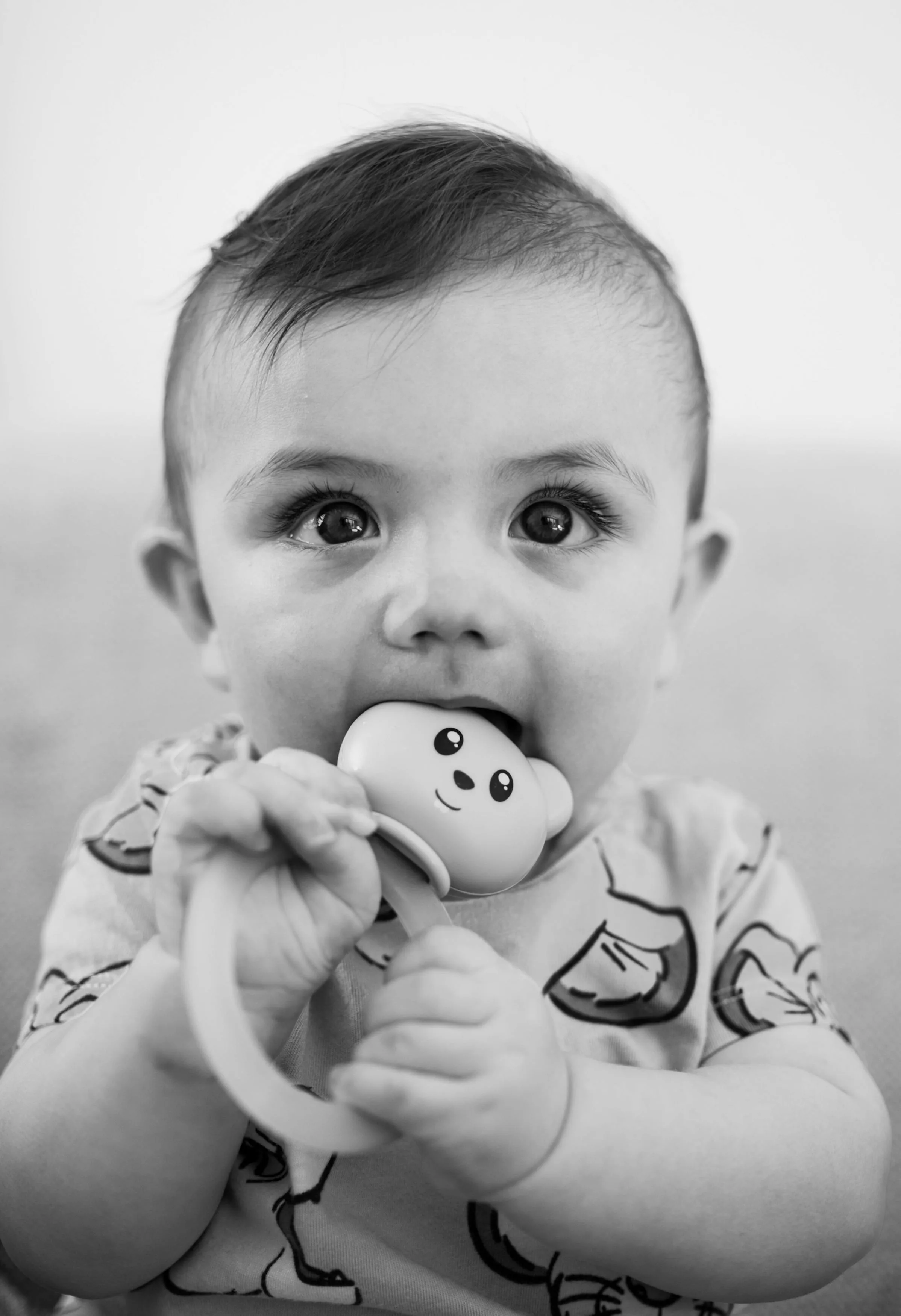 A cute baby bites a toy while sitting in a relaxed indoor setting, creating a charming portrait.