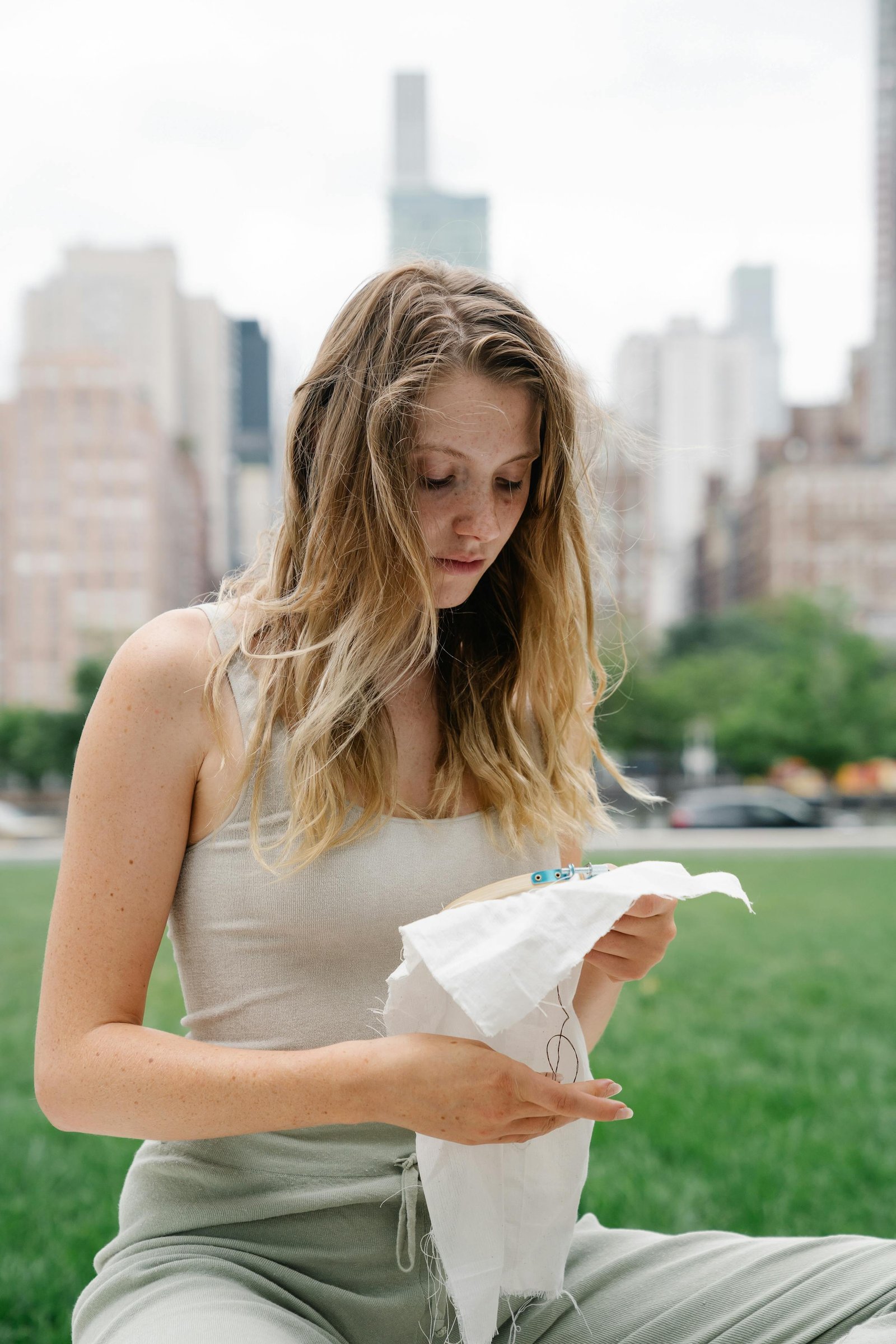 A woman sits outdoors embroidering, with a cityscape backdrop, capturing a calm urban moment.