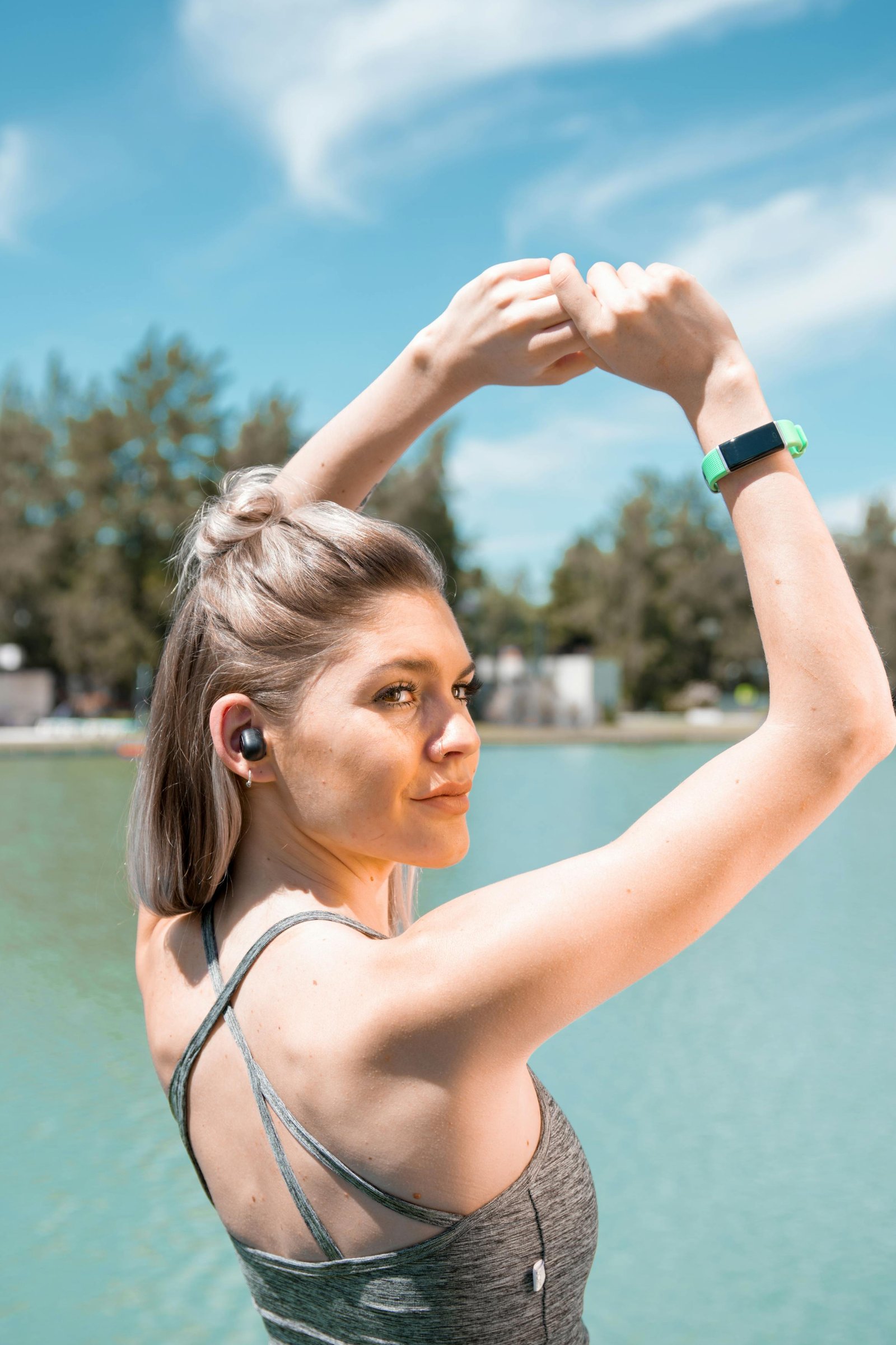 Woman stretching by a lake, wearing earphones and sportswear on a sunny day.
