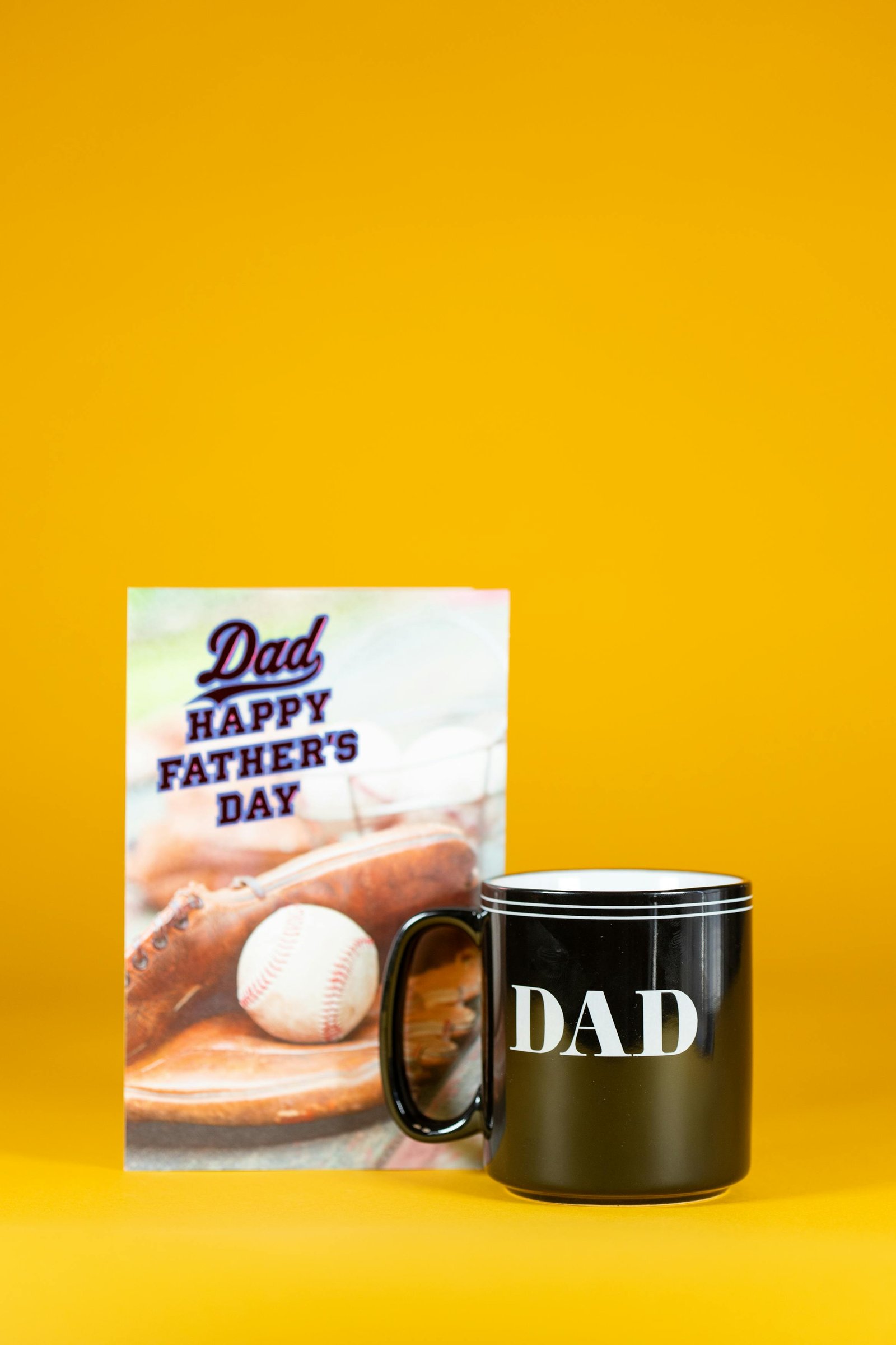 A black 'DAD' mug alongside a Father's Day card against a bright yellow backdrop.