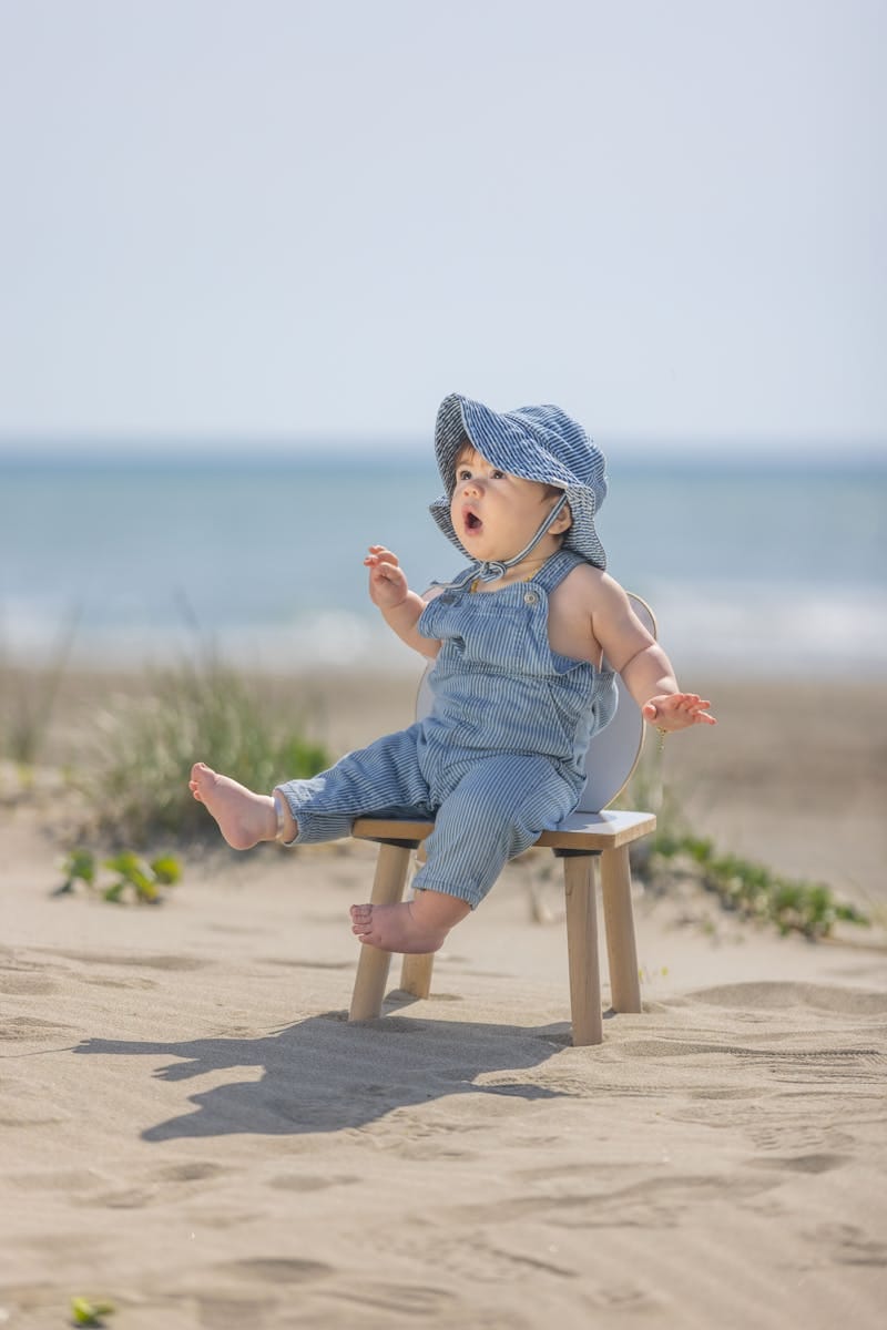 Cute baby sits on a stool at the beach, wearing blue overalls and hat.