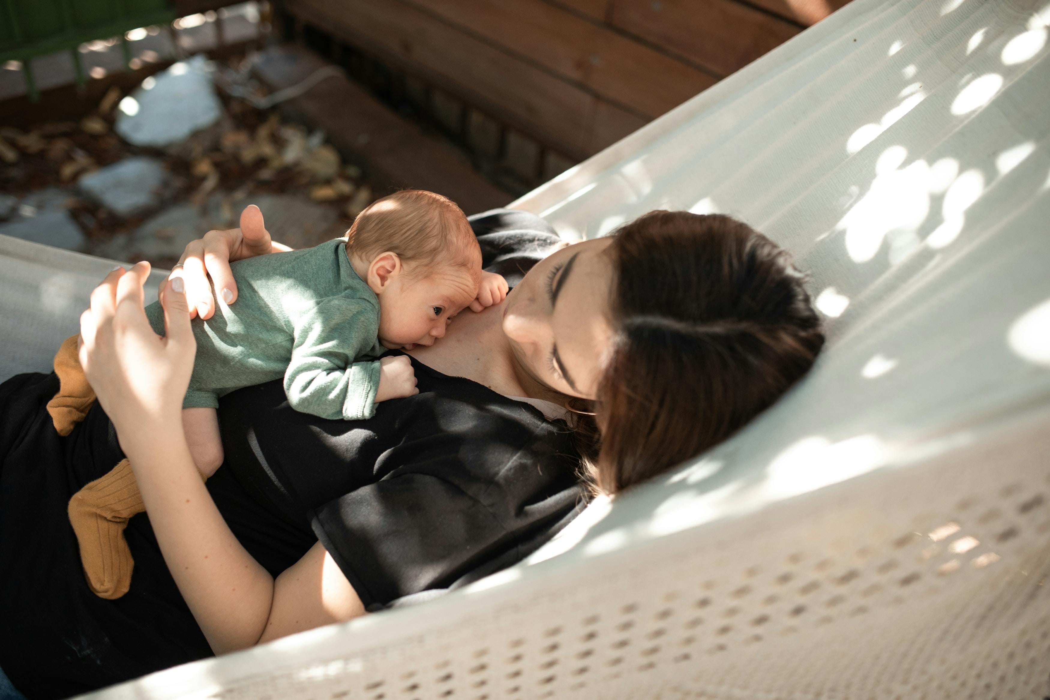 A mother gently holds her newborn baby while lying together in a hammock under sunlight.
