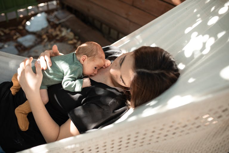 A mother gently holds her newborn baby while lying together in a hammock under sunlight.