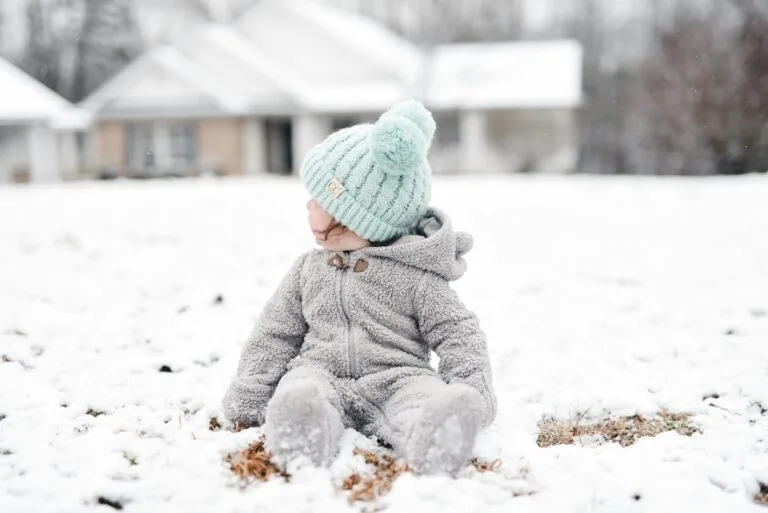 child in gray jacket and green knit cap sitting on snow covered ground during daytime